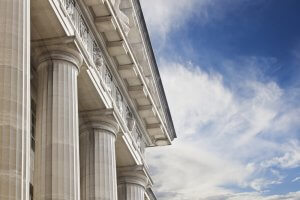 A close-up view of a neoclassical government building with large, white, fluted columns and intricate architectural details near the roofline. The sky is bright blue with scattered clouds.