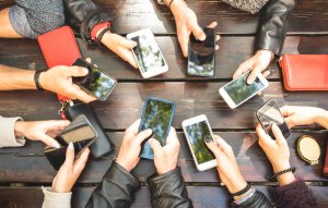 A group of people sitting around a wooden table, each holding a smartphone. Their hands, adorned with various phone cases, engage in different activities. Some red wallets and a coffee cup are also on the table, as they discuss career opportunities visible on their screens.