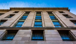 A low-angle view of a tall government building with beige stone walls and multiple large windows. The sky above is a clear blue, creating a contrast with the building's light-colored facade. The architecture features vertical lines and classical design elements.