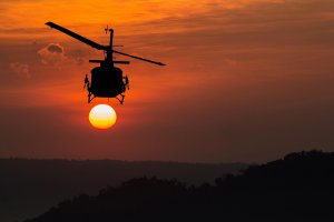 A government helicopter is silhouetted against a vibrant orange sunset. The aircraft appears to be hovering in the sky, with the setting sun directly behind it, creating a dramatic and striking visual effect. The horizon features a darkened, wooded landscape.