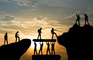 Silhouettes of people working together to build a bridge between two cliffs at sunset. Some hold and support the bridge, while others walk across. Individuals on the opposite cliff raise their hands in encouragement, symbolizing trust, teamwork, and collaboration.