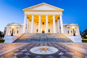 The image shows the Virginia State Capitol building in Richmond during the early evening. The neoclassical structure, a symbol of the state's history, features grand white columns and a wide staircase leading up to the entrance. In the foreground is a circular tile design on the ground.