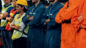 A diverse group of industrial workers in safety gear, representing their unions, stands in a line indoors. Some wear blue coveralls, one is in bright orange, and a woman wears a yellow safety vest and hard hat. They stand with arms crossed, suggesting a united, attentive stance.
