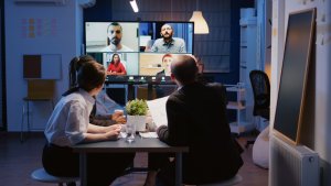 Four people sit at a table in a dimly lit office, participating in a remote video conference with five individuals displayed on a large screen. The group at the table appears engaged, discussing and pointing at the screen. A small potted plant and documents are on the table.