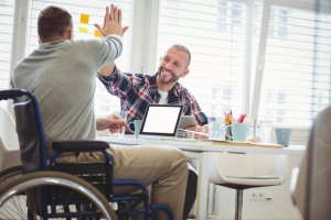 Two men in an office are celebrating a success. One is in a wheelchair and the other is seated, giving each other a high five. There is a laptop on the table, along with coffee cups and paper. Their cheerful expressions highlight that disabilities do not define their abilities and achievements.
