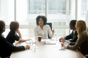 A career-driven woman is leading a business meeting in a modern office setting. She speaks while four colleagues, three women and one man, listen attentively around a conference table. Laptops, documents, and glasses of water are visible on the table.