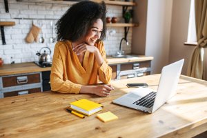 A woman with curly hair wearing a yellow blouse sits at a wooden kitchen table, smiling at a laptop during her remote work session. On the table are a smartphone, yellow notebook, and sticky notes. The background features a modern kitchen with white brick walls and wooden shelves.