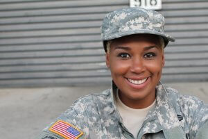 A person in military uniform smiles at the camera. The uniform features a camouflage pattern and an American flag patch on the right sleeve, embodying their commitment to government service. The individual is wearing a matching cap and is standing against a background of horizontal metal panels.