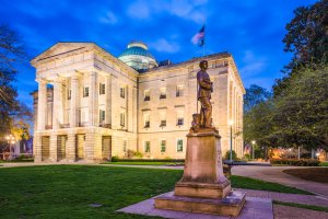 A stately, illuminated building with a dome, likely a government or historic structure, flanked by columns. A prominent statue is in the foreground set amidst a manicured lawn with surrounding trees. The twilight sky casts an enchanting glow on this symbol of the states' heritage.