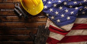 A yellow hard hat, a pair of wrenches, work gloves, and an American flag lie on a wooden surface, symbolizing the strength and unity brought by unions in the American workforce.