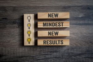 Wooden blocks on a wooden surface arranged vertically display the text "NEW MINDSET NEW RESULTS." To the left, another set of wooden blocks depicts four lightbulb icons in a vertical sequence, gradually increasing in brightness from top to bottom, symbolizing the demands of innovative thinking.