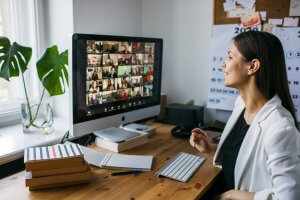 A woman in a white blazer sits at a desk with a computer screen showing a grid of people on a video call, embodying the essence of remote work. The desk has a stack of books, notepad, pen, keyboard, and mouse. A plant is near the window and a corkboard with notes fills the background.