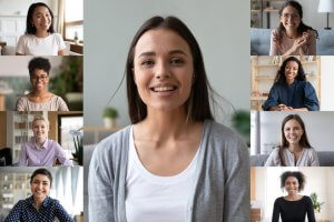 A collage of eight diverse women in separate windows surrounds a central image of a smiling woman with brown hair wearing a white shirt and grey cardigan, suggesting a remote virtual meeting or conference. All women appear to be smiling and engaged.