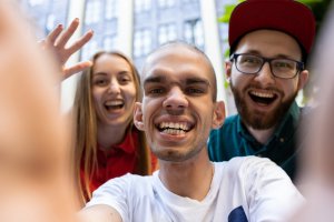 Three people are smiling and looking at the camera. The person in the foreground, who has a visible disability, is holding the camera and capturing a selfie. The group appears happy and excited, with one person waving. The background shows tall windows or buildings, indicating an urban setting.
