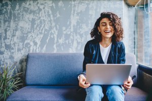 A woman with curly hair is sitting on a grey couch, holding a laptop on her lap. She is wearing a black jacket and white shirt, smiling broadly as she embraces her remote work setup. She appears to be in a relaxed, modern room with grey textured walls and a window to one side.