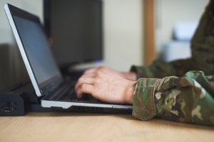 A person in camouflage military uniform is typing on a laptop. The laptop is placed on a wooden desk, and a second monitor is visible in the background. The setting appears to be indoors, possibly a government office or command center.