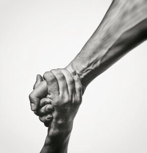 Black and white image of a strong handshake. A forearm extends from the top right to join hands firmly with another hand coming from the bottom left, symbolizing trust, support, and solidarity. The background is blurred, highlighting the hands.