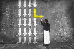 A woman stands alone facing a grungy, textured concrete wall with white arrows pointing downwards. She is drawing a large, yellow arrow to the right, diverging from the white arrows' demands. She wears a black top and white pants.