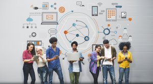 Seven people standing in a row, looking at various devices such as smartphones, tablets, and a laptop. Behind them is a grey wall with colorful icons and graphics representing remote work, social networks, cloud storage, and other digital technologies.