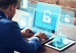 A man in a blue suit types on a laptop. Surrounding the laptop screen are floating padlock icons, symbolizing digital security. A tablet with code and password entries displayed on the screen lies next to the laptop on a wooden table. The scene emphasizes cybersecurity.