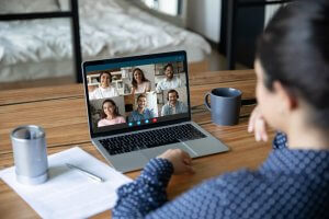 A person sits at a desk and participates in a remote video conference call on a laptop. Six people are shown in a grid format on the screen. The desk has a notebook, a pen, a coffee mug, and another cup. A bed is visible in the background.