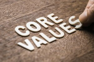 A wooden surface with the letters spelling "CORE VALUES." A hand is seen placing or adjusting the letter "S" in the word "VALUES," symbolizing leadership. The letters, made of a light-colored material, stand out against the darker wood background.