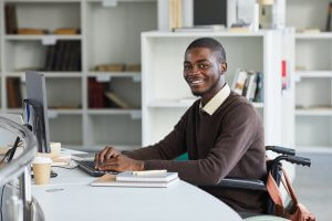 A man with disabilities sits in a wheelchair, working at a desk with a computer. He is smiling at the camera and wearing a brown sweater over a white collared shirt. The desk has notebooks, a pen, and a coffee cup, while the background features shelves with books and office supplies.