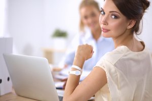 A woman with brown hair is sitting at a desk, looking back over her shoulder. She is wearing a beige blouse and a white wristwatch. In front of her is an open laptop, hinting at her busy career. Another person is sitting in the background, slightly out of focus.