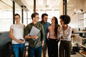 A group of five young professionals, all millennials, are standing in an office, smiling and chatting. One person holds a laptop while another cradles a coffee cup. They are casually dressed, and the office environment is modern with large glass windows and wooden accents.