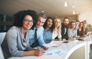 A diverse group of eight professionals, including men and women, sits at a white desk in an office setting. They are smiling and looking at the camera, with papers, graphs, and charts spread out in front of them. The room is well-lit with modern decor, reflecting their commitment to diversity.