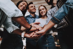 A group of five people stand in a circle with their hands stacked in the center, symbolizing unity and teamwork. Smiling in a positive, well-lit workplace environment, they are dressed casually and radiate a sense of collaboration.