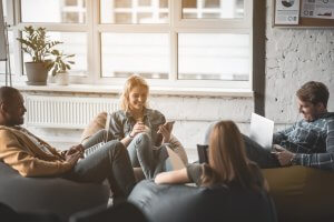 A group of four people in casual clothing are sitting on bean bags in a cozy, well-lit room with large windows. They appear relaxed, with one person using a laptop to look up government policies, another checking their phone, and others engaged in conversation. A plant is visible in the background.