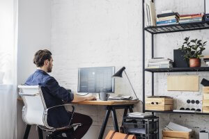 A person with dark hair is working on a computer at a desk, facing a monitor. The workspace for this remote work setup has a white brick wall, shelves with books, and various office supplies. The person is seated in a white ergonomic chair, and a desk lamp is positioned near the monitor.