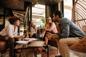 A group of four millennials, three women and one man, are sitting in a modern office lounge area, engaged in a discussion. They are seated on comfortable chairs around a coffee table with papers and pens on it. Large windows and exposed ceilings are in the background.