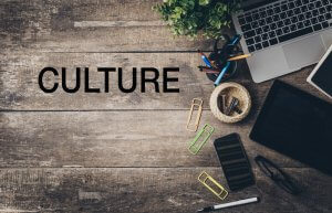 A rustic wooden desk in the workplace displays office items including a laptop, a tablet, a smartphone, colorful paper clips, pens in a cup, and a potted plant. The word "CULTURE" is prominently written on the left side of the desk.