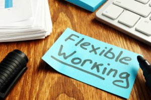 A blue sticky note with "Flexible Working" written on it is placed on a wooden desk next to a black marker, a stack of papers, and a computer keyboard, reflecting government initiatives encouraging adaptable work environments.