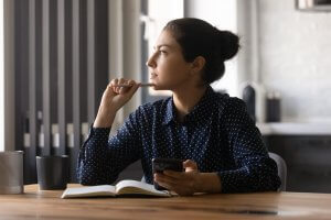 A person with their hair in a bun sits at a table, holding a pen near their mouth while looking thoughtfully into the distance. They have an open notebook and a smartphone in front of them on the table, surrounded by the sleek lines of a modern, minimalistic kitchen that serves almost like a silent servant.