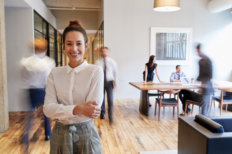 A smiling woman stands in the foreground of a modern workplace with wood floors and glass-walled rooms. In the background, people are walking around and engaged in discussions, with two individuals sitting at a conference table. The atmosphere is busy and professional.