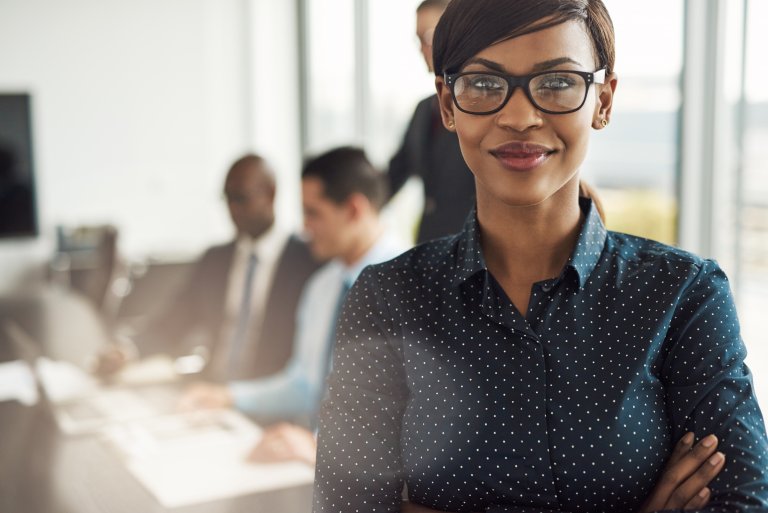 A confident woman with short hair wearing glasses and a navy blue polka-dot shirt stands in the foreground with her arms crossed, embodying leadership. In the background, three men are sitting and discussing in a bright, modern office.
