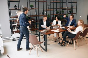 A group of five people is in a modern meeting room. One person stands near a whiteboard, presenting to four seated colleagues who are listening attentively and honing their soft skills. The room has a large wooden table, scattered papers, laptops, and a large shelving unit with various items.