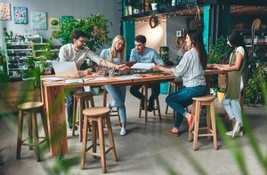 A group of five people work collaboratively around a wooden table in a modern, well-lit workplace with plants and art on the walls. They are discussing papers and using laptops. Some are seated on bar stools while others stand or lean on the table.