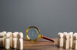 A magnifying glass with a gold rim and wooden handle is placed on a wooden surface. Positioned between two groups of small wooden human figures facing each other, it symbolizes the scrutiny or investigation to reveal the truth between two parties. The background is plain grey.