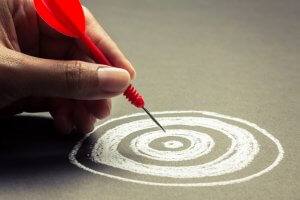 A hand, like a loyal servant, holds a red dart poised above a chalk-drawn target on a blackboard. The target consists of concentric circles, with the dart aiming toward the center.
