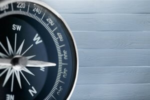 Close-up of a black and silver compass symbolizing leadership, showing cardinal directions W (west) and S (south), positioned on the left side of the image. The background consists of light gray wooden planks lined horizontally.