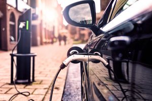 A close-up view of an electric car being charged at a station on a bustling city street. The charging cable, plugged into the side of the electric vehicle, reflects the sun's warm light, while tall buildings loom in the background.