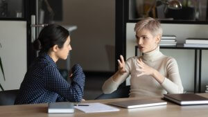 Two women are seated at a desk, engaged in a serious conversation about their careers. One woman with short blonde hair and a white turtleneck is gesturing with her hands, while the other woman with dark hair, tied back, is listening attentively. Laptops and documents are on the desk.