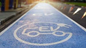 A blue road lane with a white electric vehicle (EV) icon and plug symbol painted on it, designating it for electric cars. The electric path stretches into the distance, illuminated by a warm light.