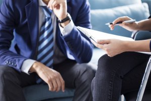 Two individuals are engaged in a business meeting. One holds a clipboard and pen, preparing to write, while the other, dressed in a suit and tie, is seated and appears to be listening intently with one hand on his chin. Both are partially visible as they discuss career opportunities.