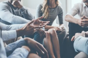 A group of people sitting closely together in a casual setting, engaged in conversation. The focus is on their hands and gestures, emphasizing active participation and leadership. The individuals are dressed in casual clothing, indicating a relaxed and informal meeting.