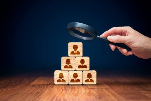 A hand holds a magnifying glass over a pyramid of wooden blocks on a wooden surface. Each block features an engraved silhouette of a person, representing recruiting or employee selection. The background is dark blue, accentuating the blocks in the foreground.